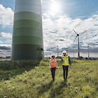 An engineer and a DEKRA engineer are walking across a meadow. Several wind turbines can be seen behind them.