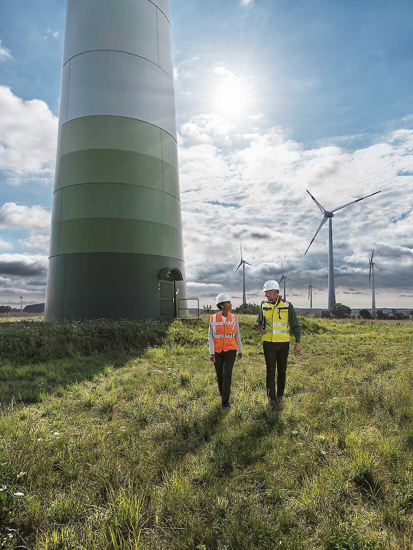 Eine Ingenieurin und ein DEKRA Ingenieur laufen über eine Wiese. Hinter ihnen sind mehrere Windräder zu sehen.