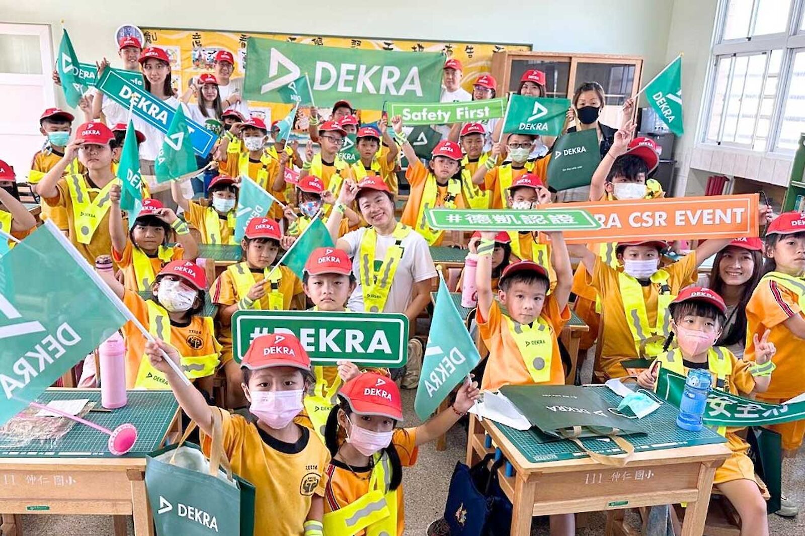 A classroom with lots of children wearing DEKRA children's caps and holding shields with DEKRA written on them.